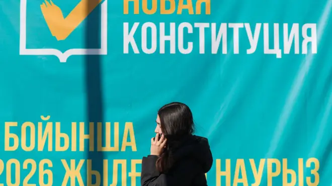 Девушка в темном пальто с непокрытой головой говорит по телефону, проходя мимо плаката, призывающего голосовать на референдуме о новой Конституции Казахстана