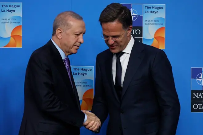 Erdoğan ve Rutte el sıkışıyor. Arkalarında mavi renkli bir pano var. Panonun üzerinde NATO amblemi ve zirvenin posteri bulunuyor. 