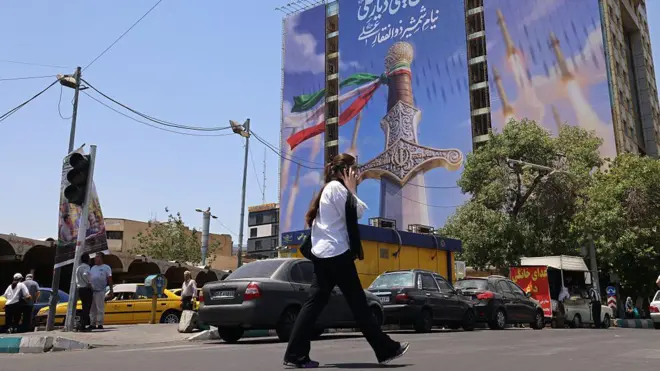 امرأة تمر أمام لافتة عملاقة تصور صواريخ إيرانية وسيفًا للإمام علي، الإمام الأول للشيعة، في ساحة فاناك في طهران في 10 يونيو/حزيران 2026. 