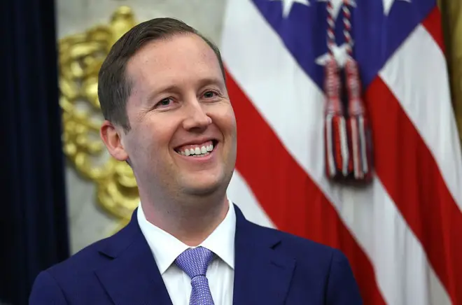 Сержио Гор мовий костюм, оқ куйлак устидан мовий галстук таққан, камерага табассум қилиб қараяпти, орқасида АҚШ байроғи