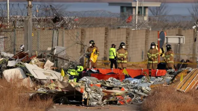 Bombeiros, algunsbet35trajes marrons e outrosbet35branco e preto, vasculham os destroços da aeronave Jeju Air. Os destroços são quase identificáveis ​​como um avião, exceto pela cauda