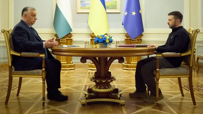 Зеленський у чорному светрі та військових черевиках (праворуч) і Орбан у темному костюмі (ліворуч) сидять один навпроти одного за круглим дерев’яним столом. Між ними на столі стоїть букет синьо-жовтих квітів, а позаду – три прапори: Угорщини, України та Європейського Союзу