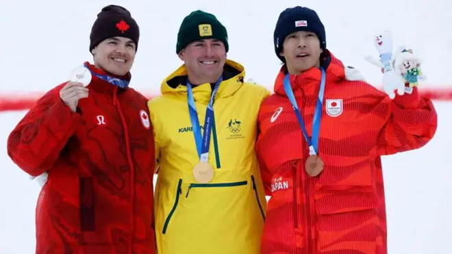 3人が首にメダルをかけて横並びに立ち斜め前方を向いている。堀島は大会マスコットのぬいぐるみを持った左手を上げている