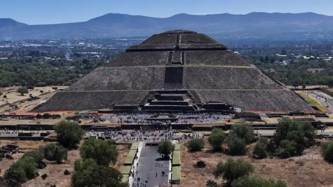 A big pyramid seen from a distance with mountains in the background, and groups of people in the foreground