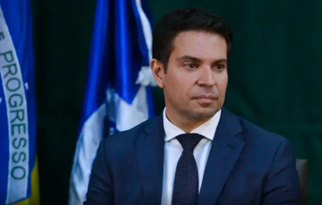 Foto de Alexandre Ramagem usando terno diante de uma bandeira brasileira e olhando para a direita em direção ao vazio com aparência distante