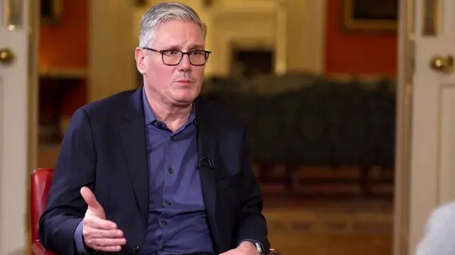 Keir Starmer, mavi gömlek ve koyu renk ceket giymiş halde kırmızı bir sandalyede otururken el hareketleri yapıyor.
