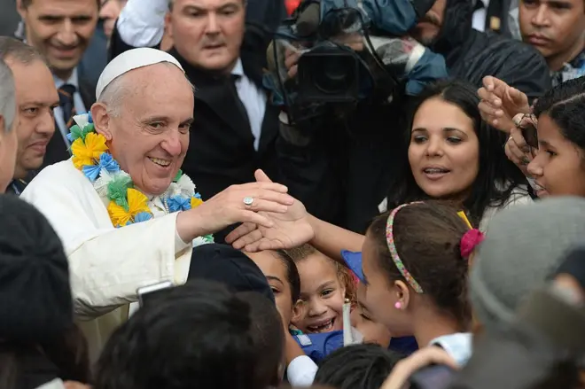 Papa Francisco sorri enquanto cumprimenta crianças e mulheres; ele usa um colar amarelo, verde, azul e branco