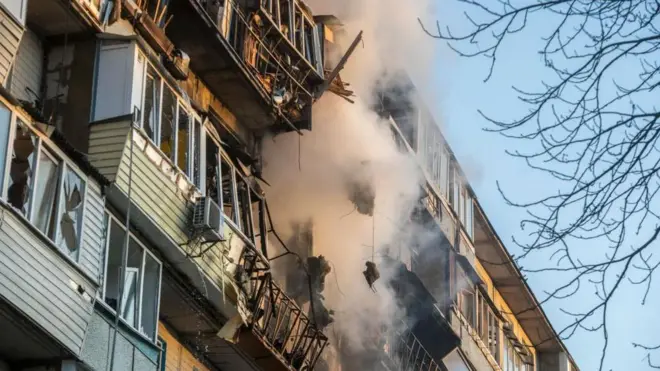 青空を背に、多層階の集合住宅の高層階の窓が焼けこげ、白い煙が出ている。その周りの窓や壁も破損している