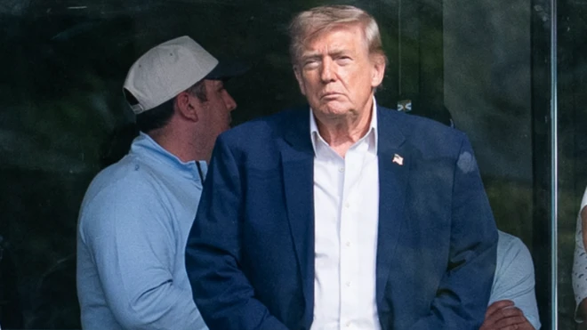Trump elleri kemerinde bir şekilde uzaklara bakarak bir şeyler izliyor.