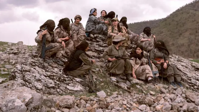 Female PJAK fighters holding their weapons sit on a rocky hilltop. Some wear head and face coverings and many carry weapons.