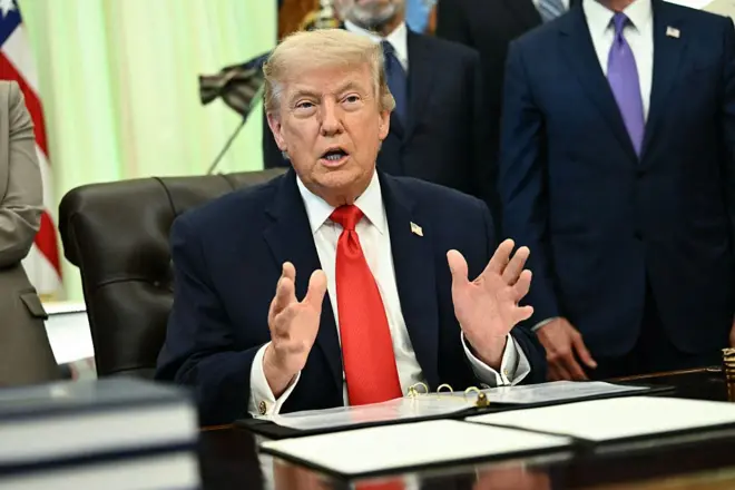الرئيس الأمريكي، دونالد ترامب، مرتديا بدلة زرقاء وقميصا أبيض ورابطة عنق حمراء، ويتحدث وهو جالس داخل المكتب البيضاوي في البيت الأبيض، وخلفه عدد من المسؤولين الأمريكيين.