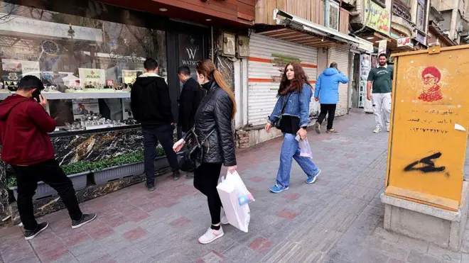 چند زن و مرد در حال عبور از مقابل یک مغازه ساعت‌فروشی با تصویری از علی خامنه‌ای، رهبر کشته‌شده ایران، روی یک ورق فلزی زرد رنگ در میدان تجریش تهران. 