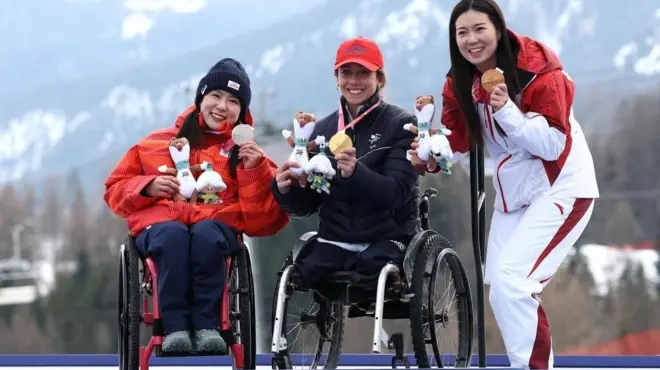 3選手が表彰台の上で横に並んでいる。全員笑顔で、左手でメダルを持ち、胸の前で見せている。背後には山並みが見える