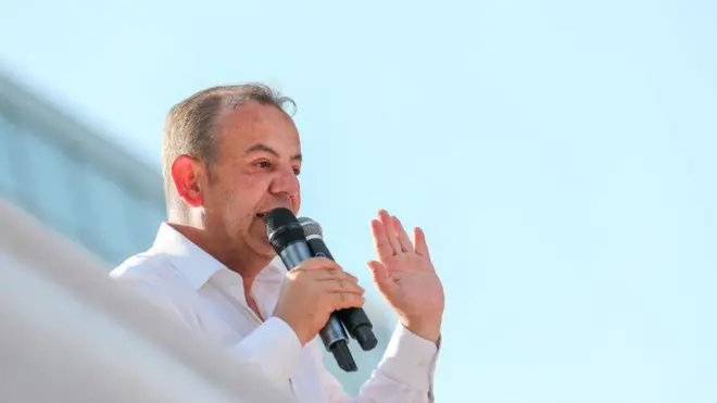 Tanju Özcan üzerinde beyaz bir gömlek ve elinde iki mikrofonla bir konuşma yaparken