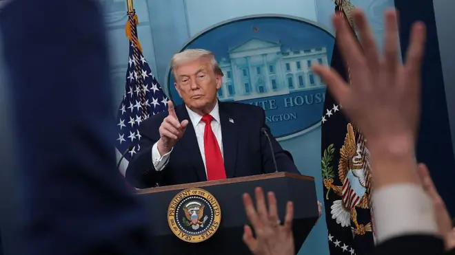 Donald Trump, soru sormak için el kaldıran gazetecilerin ellerinin arasından Beyaz Saray'ın kürsüsünde görülüyor. Başparmağını söz vereceği gazeteye uzatmak üzere kaldırmış