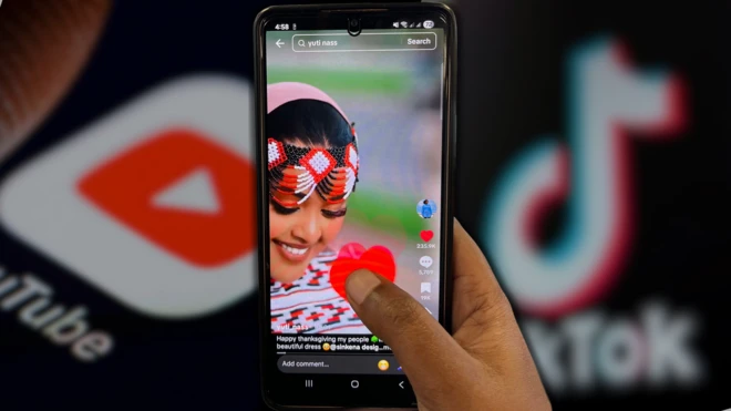 ቲክቶክ የተከፈተበትን እጅ ከያዘ ስልክ ጀርባ የዩቲዩብ እና ቲክቶክ ሎጎዎች ይታያሉ