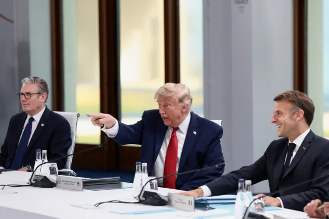 Keir Starmer brit miniszterelnök, Donald Trump amerikai elnök és Emmanuel Macron francia elnök egy fehér asztalnál ülnek. Donald Trump jobb kezét felemeli, és előre felé mutat.