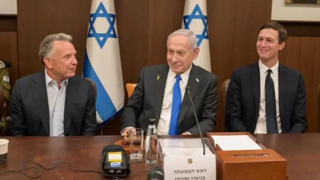 Netanyahu entre dois homens diante de mesa, com bandeiras de Israel ao fundo; eles riem