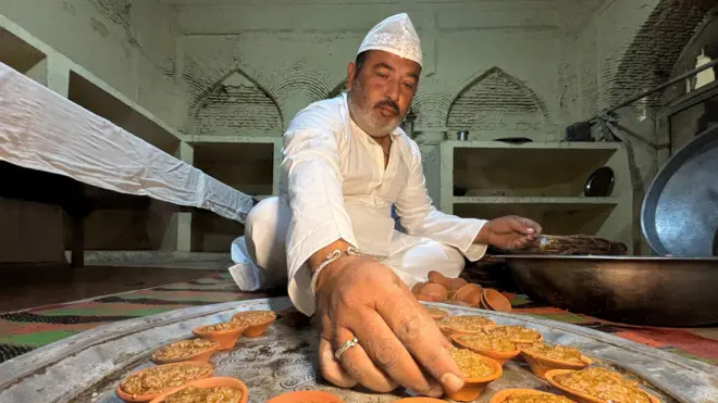 Murtuza Hussain Raju, wearing a white kurta and a skull cap, prepares kebabs while sitting on the floor. 