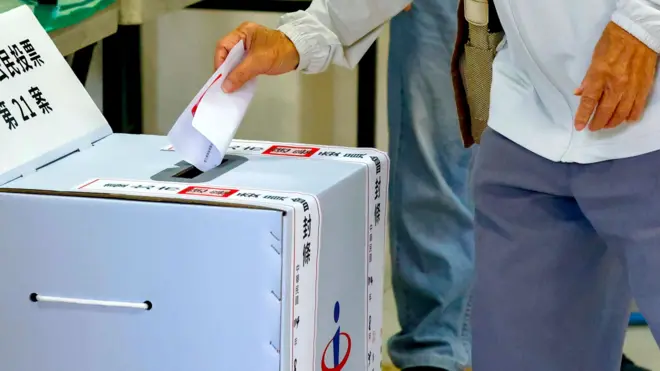 台北某投票所內一位身穿灰色外套的老人將「重啟核三」公投票投入貼上了封條的紙板票箱（23/8/2025）