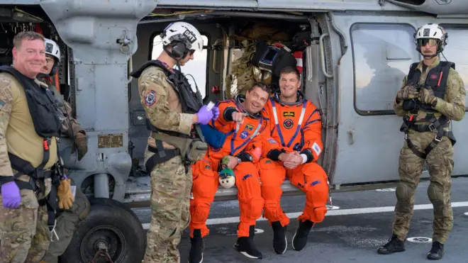 Dva astronauta u narandžastim odelima sede na dovratku helikoptera, a oko njih je nekoliko vojnika