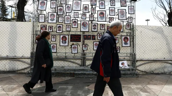 Tahran'da sokakta yürüyen bir kadın ve erkek, arka planda, savaşta ölenlerin fotoğraflarından oluşan bir duvar panosu var
