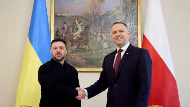У приміщенні Зеленський і Навроцький тиснуть руки, стоячи між українським та польським прапорами. На стіні за ними висить велика картина з історичним сюжетом.