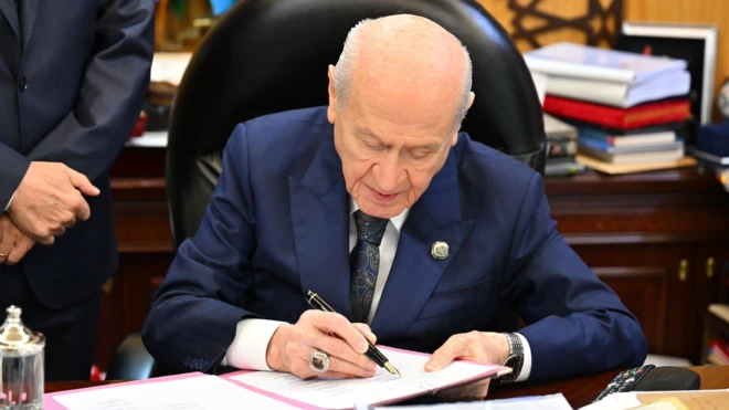MHP Genel Başkanı Devlet Bahçeli, önündeki kağıtlara imza atıyor. Arkada ellerini önden bağlamış bir kişi görülüyor.