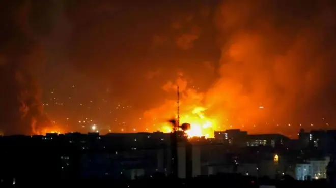 暗い建物群の向こうでオレンジ色の大きな火の手が複数上がり、煙が夜空に立ち上っている
