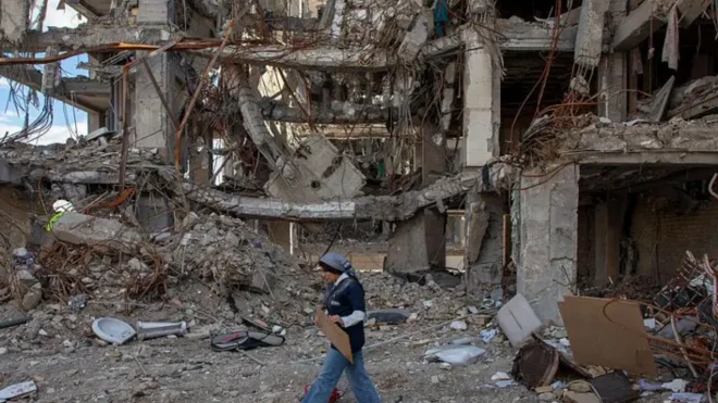 زنی با لباس آبی و شلوار جین در کنار یک ساختمان تخریب شده 