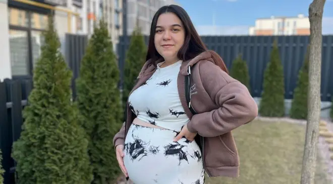 Uma mulher grávida está em pé em um jardim com a mão sobre a barriga. Ela tem cabelo longo, liso e escuro e veste um conjunto de blusa e calça brancas com manchas pretas. Também usa um moletom marrom-claro