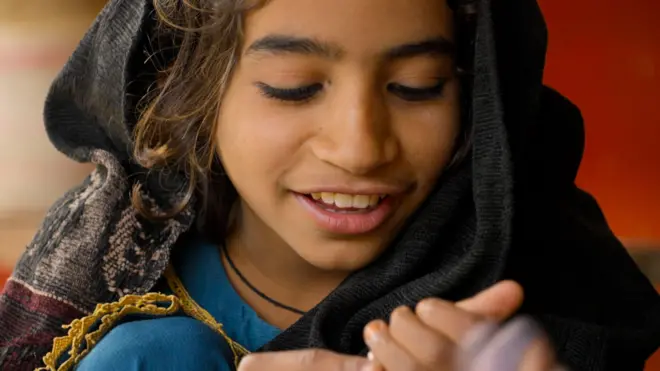 Una niña con un vestido azul sonríe, con la mirada fija en sus manos. Lleva un pañuelo oscuro con un borde estampado sobre la cabeza. 