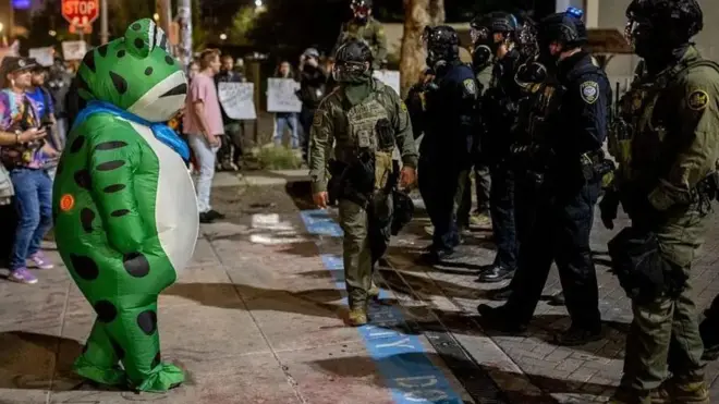 در یک تجمع که معترضان مقابل صف پلیس ایستادهاند، فردی در لباس قورباغهای در جلوی صف معترضان و مقابل پلیس ایستاده است