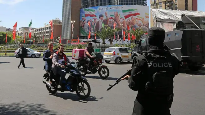 یکی از میدانهای تهران در حالیکه مامور پلیسی که صورت خود را پوشانده و اسلحه در دست دارد ایستاده. عابران و دو موتور سوار در حال گذار از خیابان هستند.