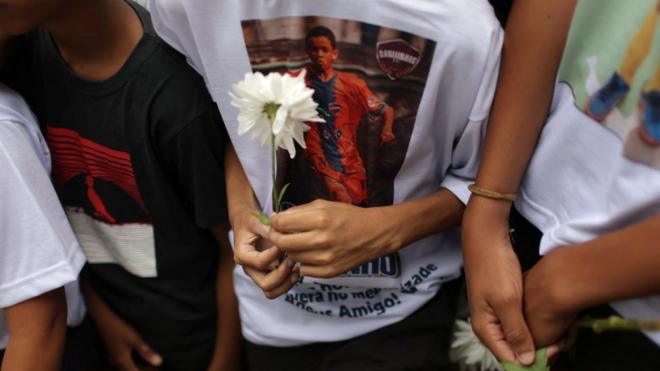 Três adolescentes em enterro, com flores na mão e um deles com camisa estampando foto de Thiago Menezes Flausino