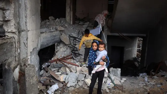 طفلة ترتدي حجاباً تحمل طفلة أخرى وخلفها طفل وامرأة يقفان على الأنقاض