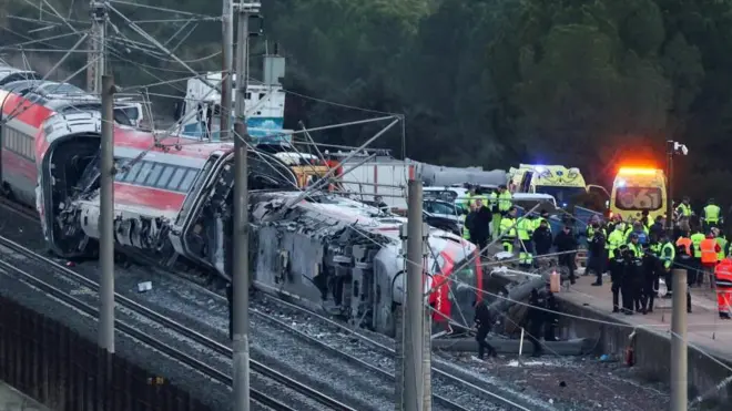 赤い列車が数両、脱線して横倒しになったり、傾いたりしている。付近には治安警備隊などの当局者らが多数いる