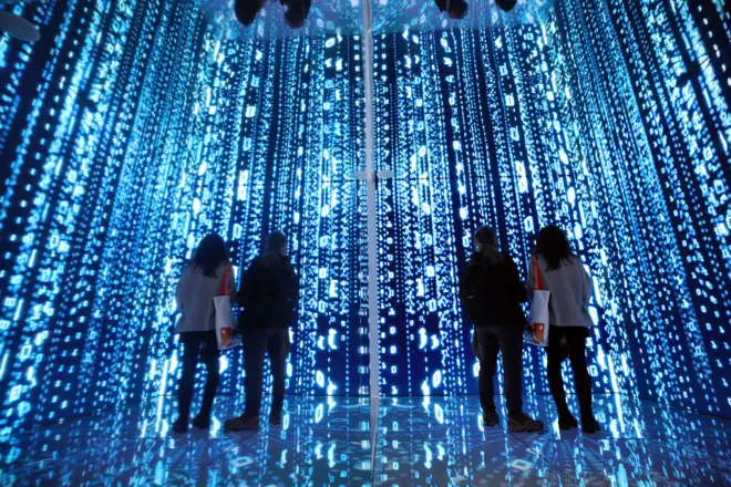 파란색 빛의 디지털 공간에서 밝은 숫자와 기호들이 벽과 바닥을 가득 채우고 있으며 관람객이 미디어 아트를 감상하고 있다