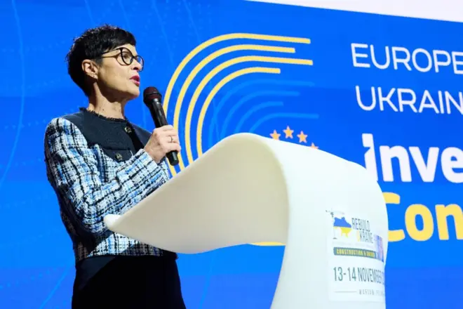 Еврокомиссар по вопросам расширения Марта Кос в синем платье с клетчатыми рукавами с микрофоном в руках выступает с белой трибуны на фоне бело-желто-голубого стенда, на нем справа части слов "Европа" и "Украина"