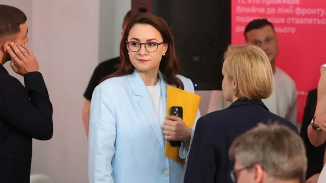 Yulia Svyrydenko, dressed in a light-blue suit, holds a yellow folder and mobile phone. She's surrounded by other people