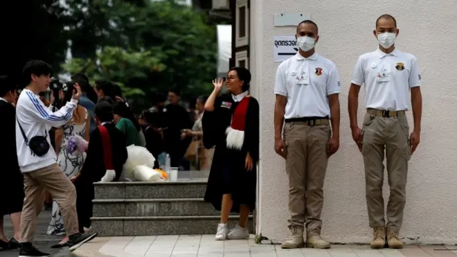 บัณฑิตถ่ายรูปข้างหอประชุม ขณะที่เจ้าหน้าที่ยืนรักษาความปลอดภัย