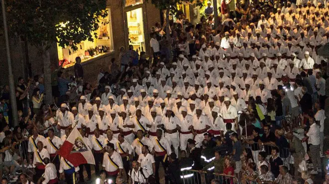 Bir geçit töreninde beyaz kıyafetler giymiş çok sayıda kişi yan yana yürüyor. 12 kişiden oluşan sıralarda insanlar kol kola girmiş ve art arda onlarca sıra görülüyor