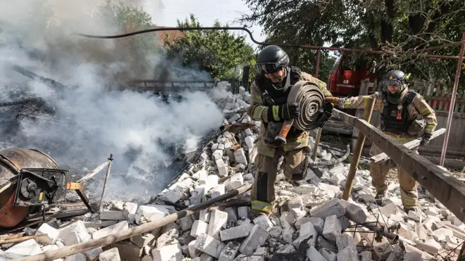Two firefighters in full gear stand on burning rubble while holding a rolled up hosepipe, about to release water to battle the flames.