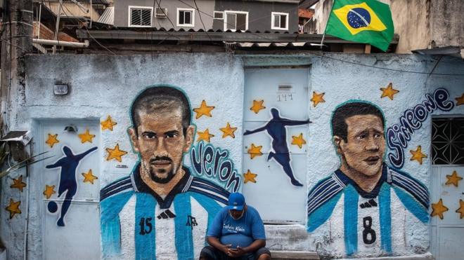 Homem sentado no meio fio, em frente a muro grafitado com imagens de dois jogadores de futebol argentinos; em cima do muro, há uma bandeira do Brasil