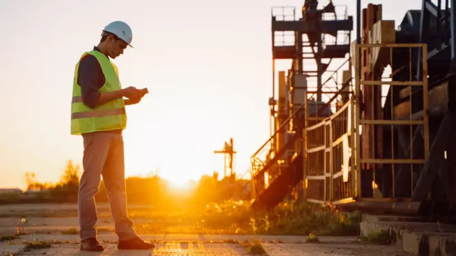 Un hombre con chaqueta de alta visibilidad y casco, midiendo y analizando datos de un campo petrolífero cerca de una construcción 