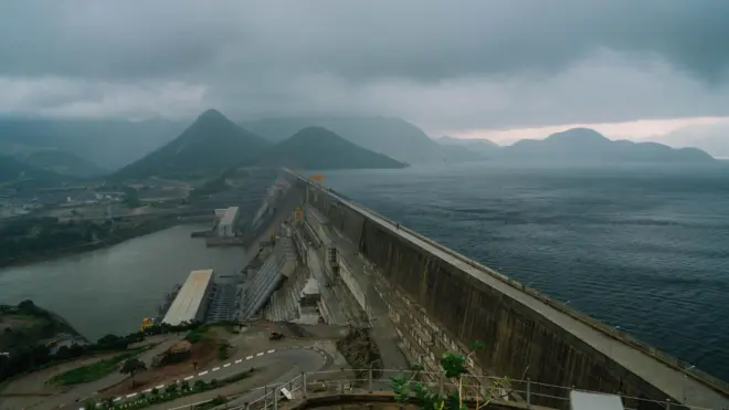 The completed dam. On one side of the wall is a vast lake and on the other side is industrial infrastructure.