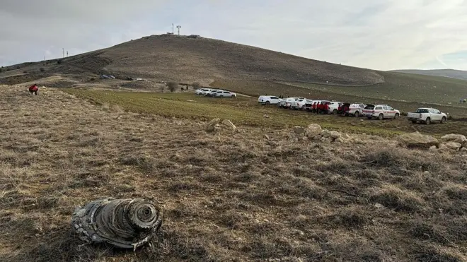 Uçağın enkazından bir parça bir tarlada görünüyor ve arka plandaki tepenin alt kısmında araçlar park etmiş halde duruyor