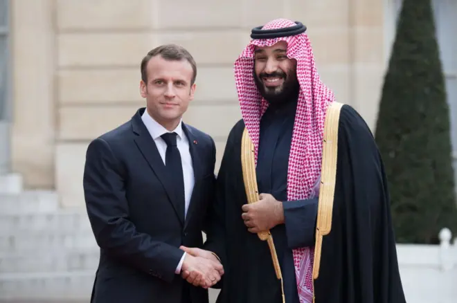 بن سلمان يرتدي ملابس السعودية التقليدية بلون أسود وبجانبه ماكرون ببدلة كحلية رسمية مع ربطة عنق بنفس اللون وقميص أبيض.