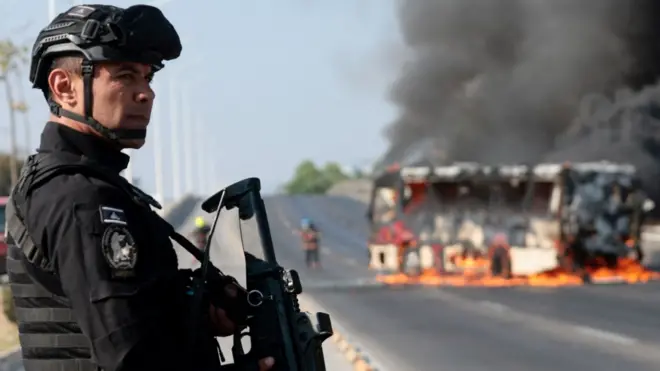 A member of the Prosecutor's Office holding a machine gun in front of a burning bus in Zapopan, Jalisco state, Mexico, on 22 February 2026. Photo by Ulises Ruiz.
