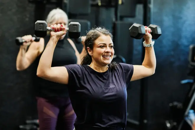 Strock fotoğrafı, sağ omzunda çiçek dövmesi olan bir kişiyi spor salonu ortamında yanal ağırlık çubuğunu aşağı çekerek kırmızı ve pembe spor kıyafetleri giyen gösteriyor..